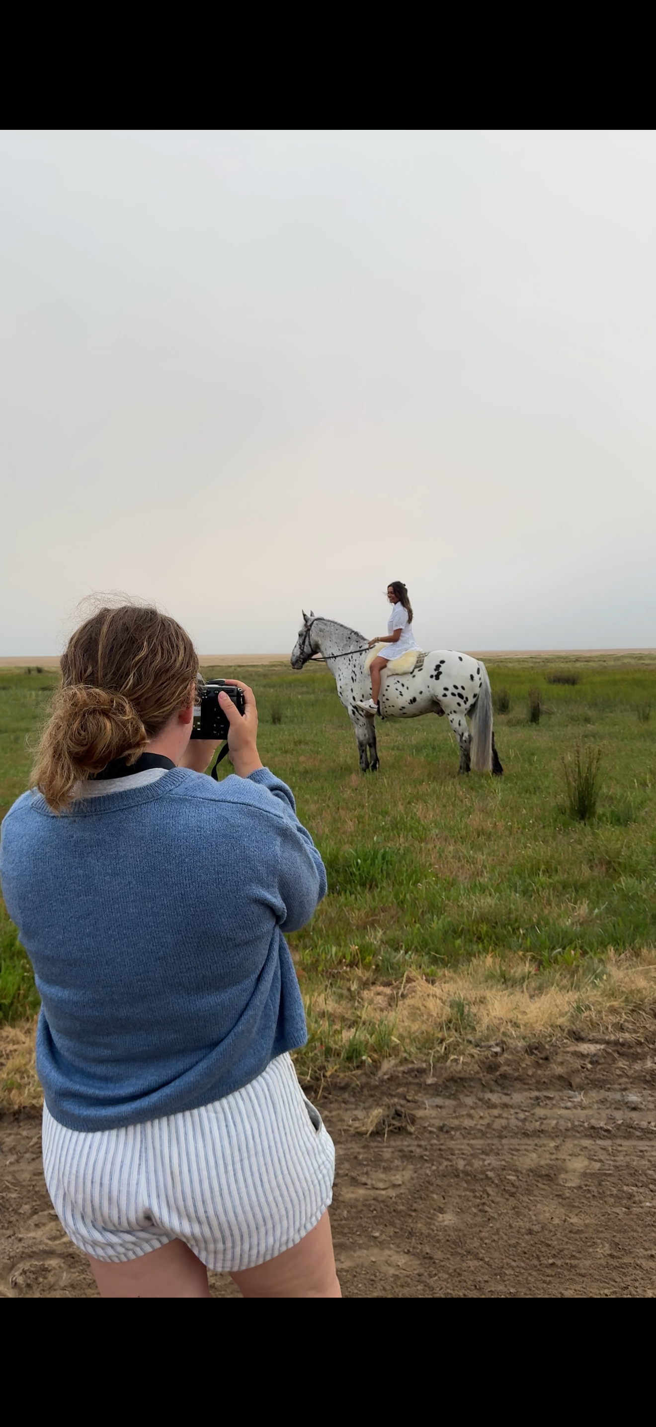 A woman in a blue sweater photographs a rider on a spotted Appaloosa horse in a vast green meadow under a hazy sky.