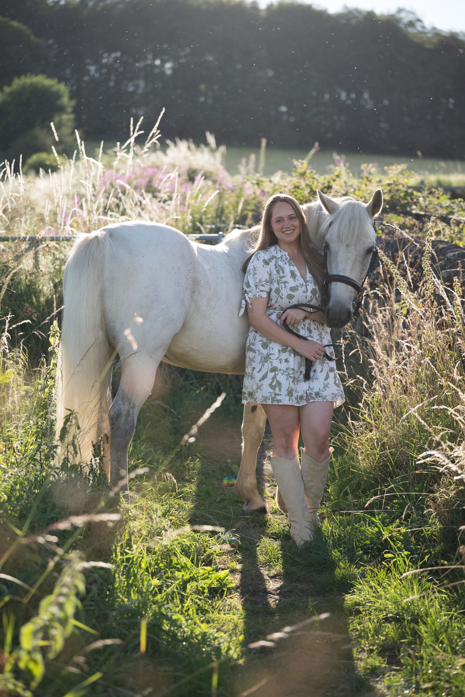 A young woman in a floral dress stands with a white horse in a sunlit green field.