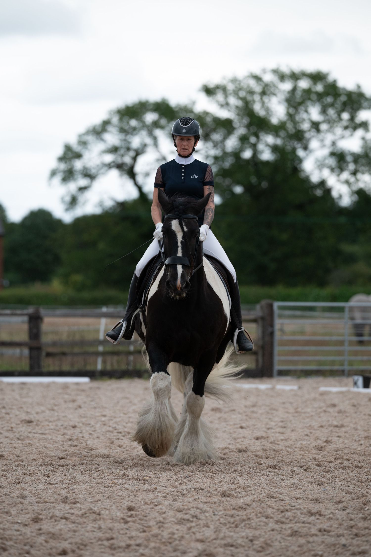 A rider in black attire and helmet rides a black and white Gypsy Cob in a sandy arena.