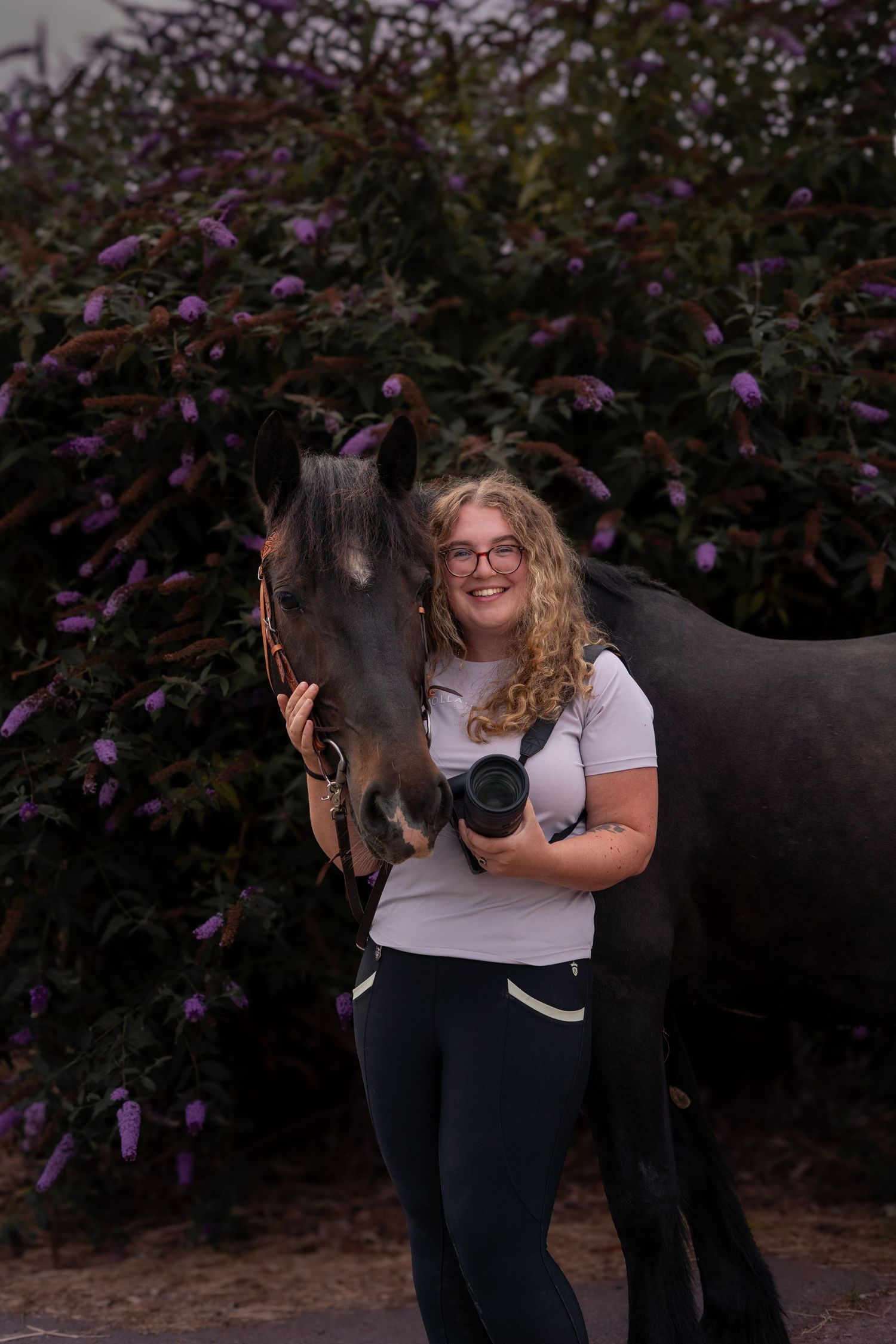 Smiling blonde woman with glasses holds camera beside dark horse in front of purple flowering bushes.