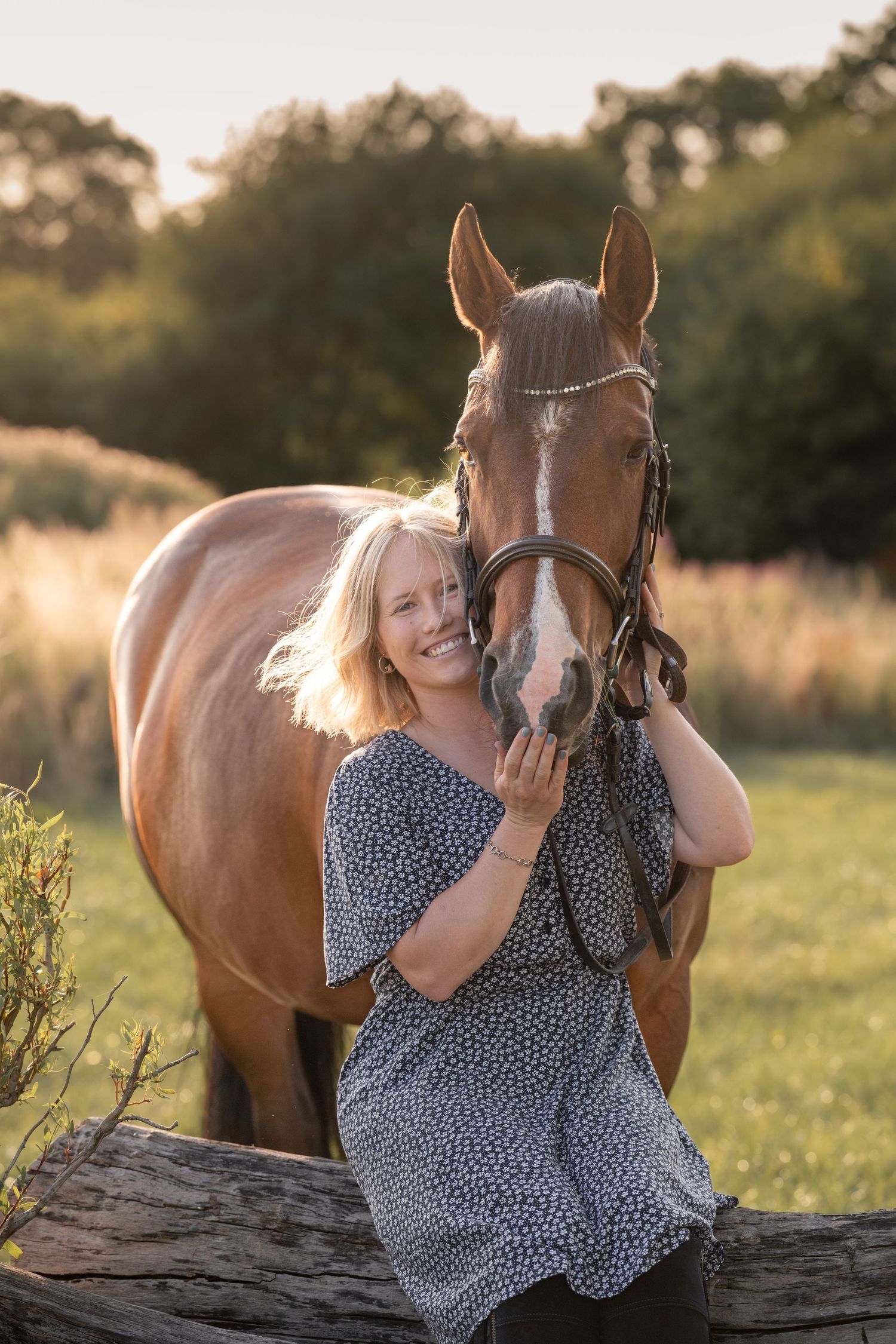 Smiling blonde woman in floral dress embracing a chestnut horse in a sunlit field at golden hour.