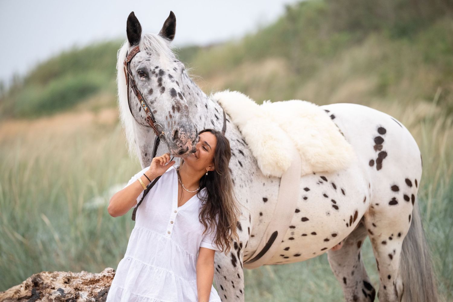 Woman in white dress affectionately nuzzling an Appaloosa horse in a scenic outdoor field setting.