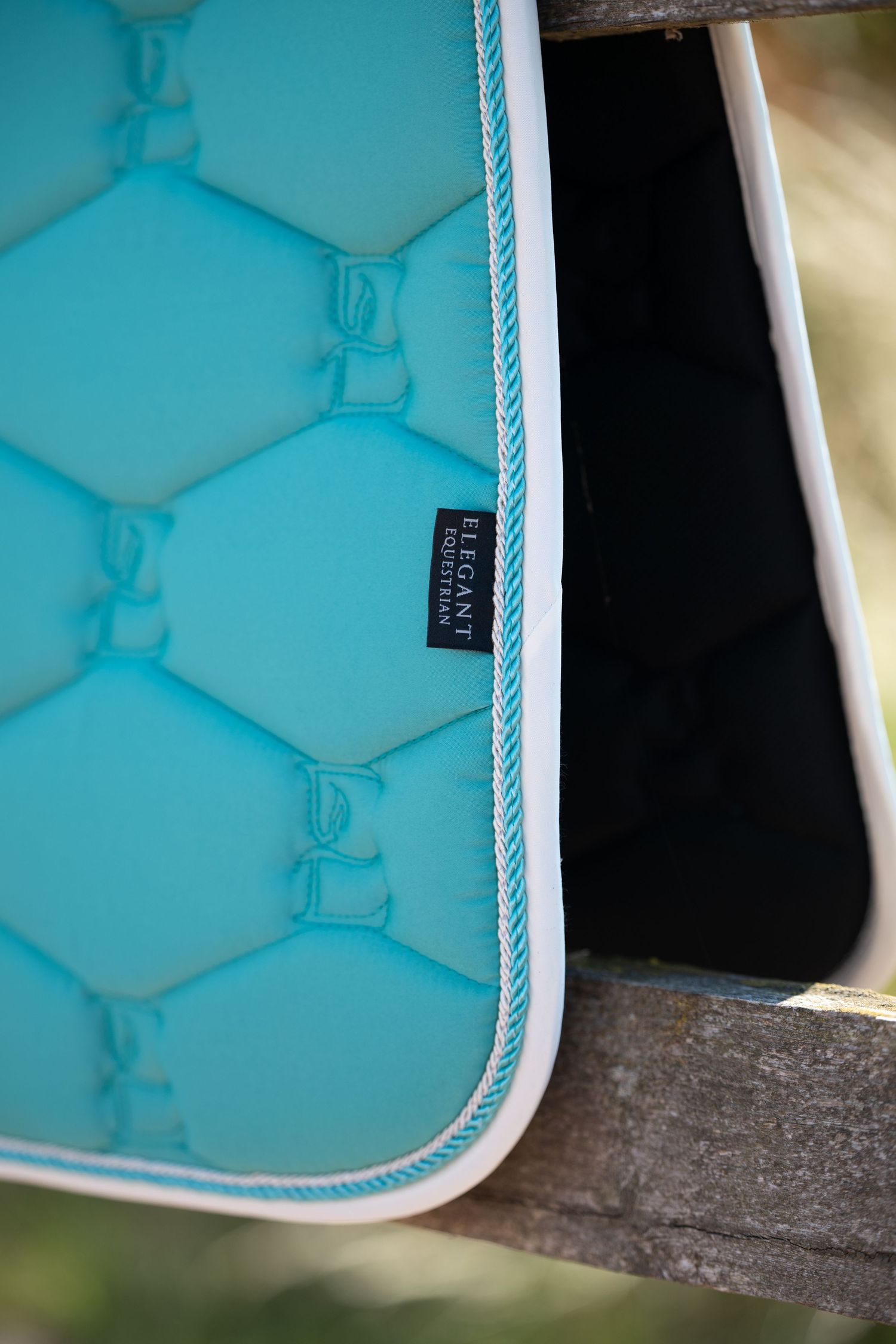 Turquoise quilted horse saddle pad with white trim and Elegant Equestrian brand label on wooden fence.