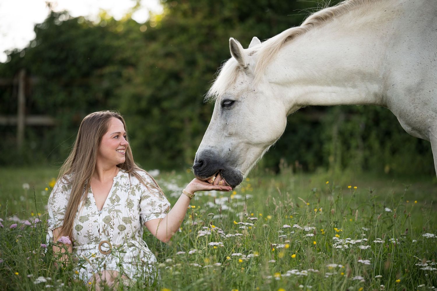 A smiling woman feeds a white horse by hand while sitting in a daisy-filled meadow.