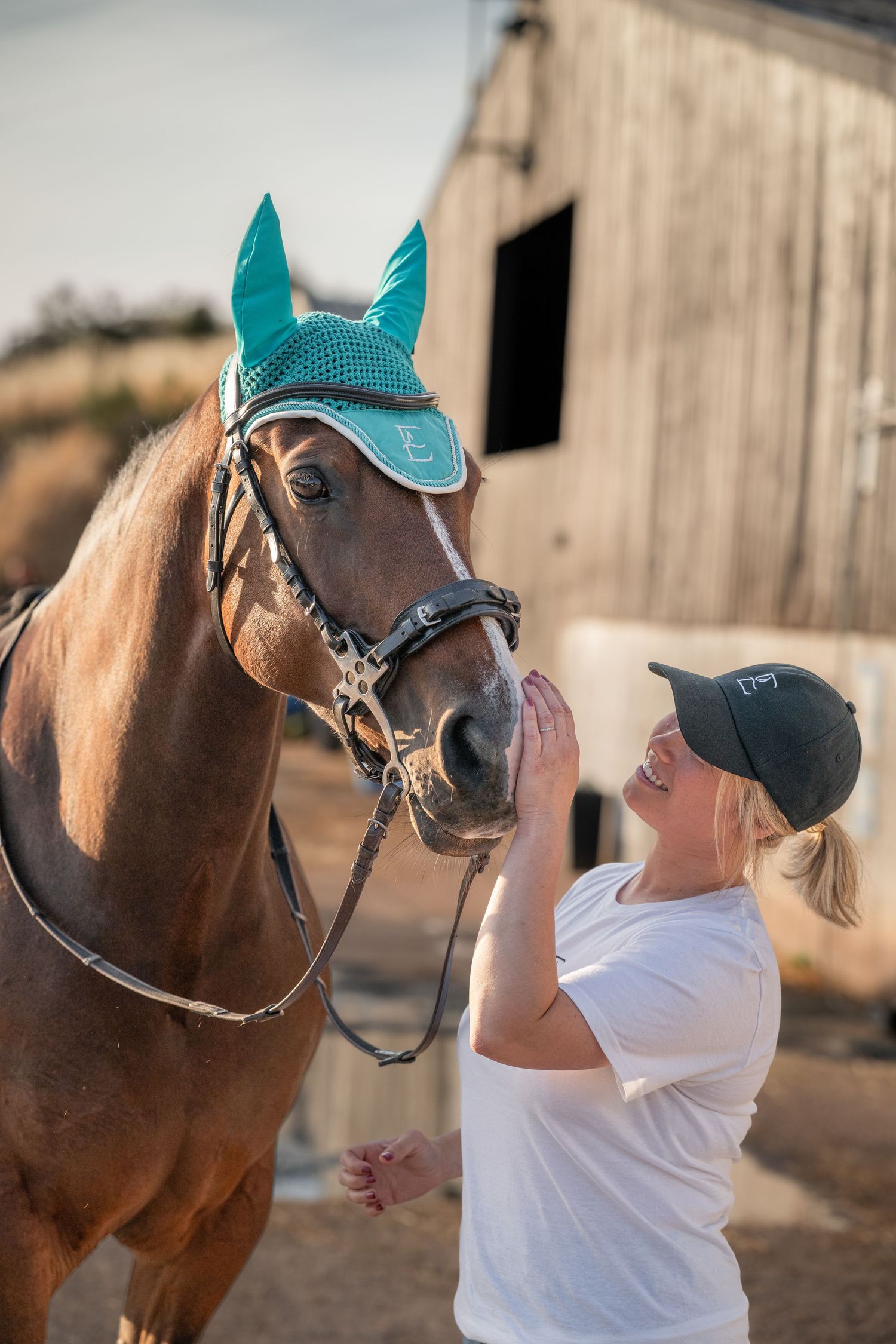 Smiling blonde woman in white shirt and cap interacting with a chestnut horse wearing a teal bonnet near a barn.