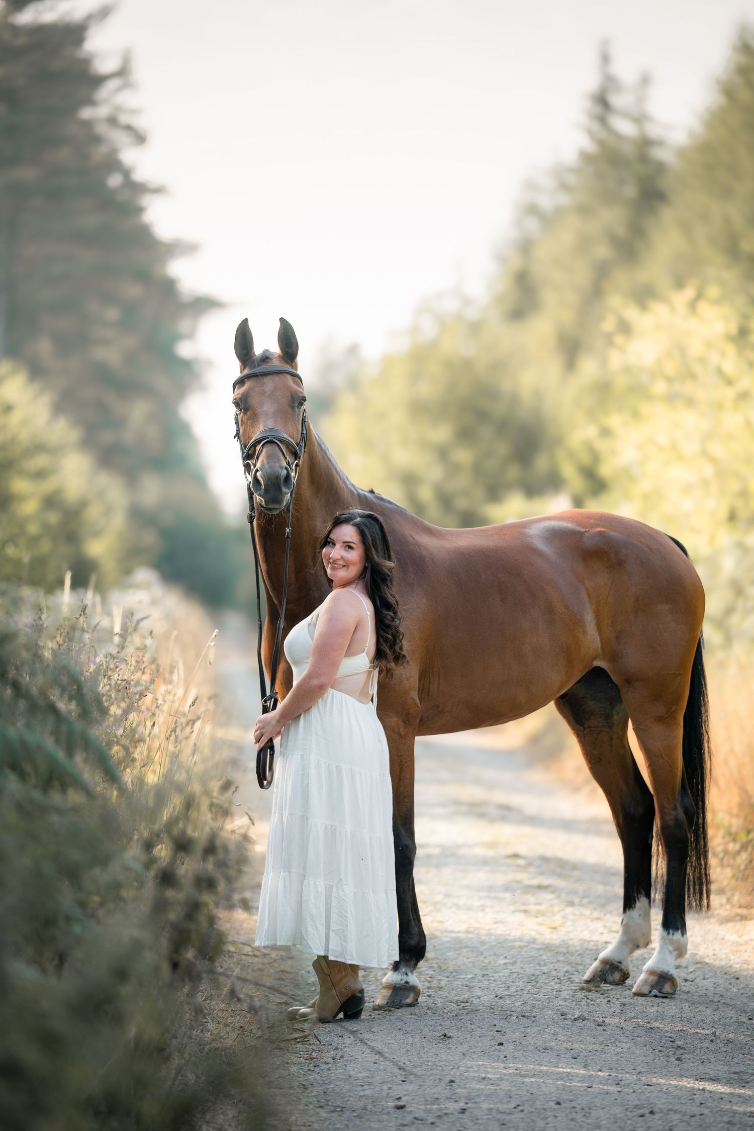 Woman in white dress standing beside a bay horse on a tree-lined path in soft golden light.