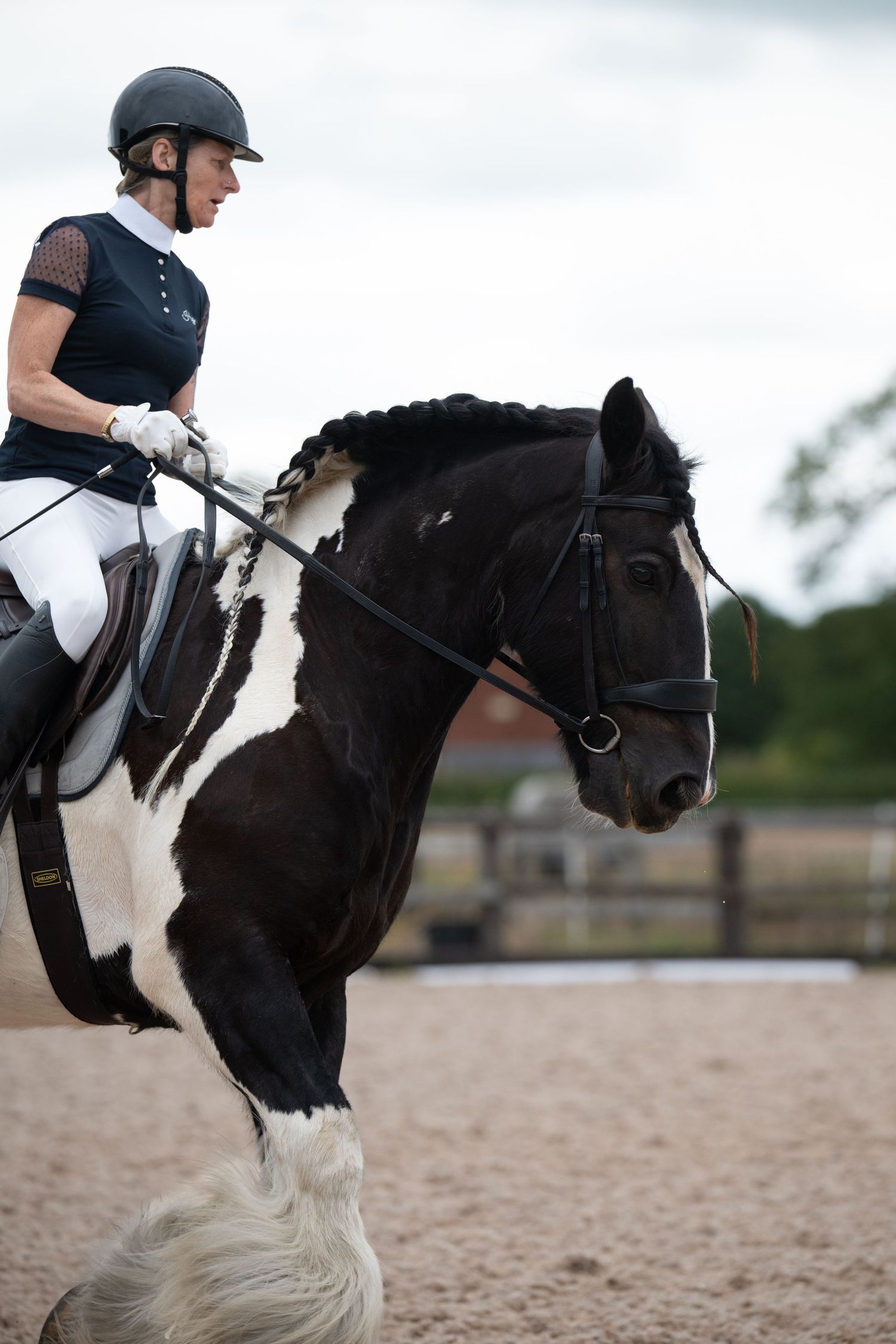 Female rider in dressage attire riding black and white pinto horse in sand arena.