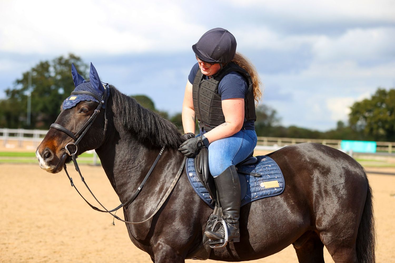 Woman riding a dark bay horse in an outdoor arena, wearing a navy helmet and blue jeans during an equestrian session.