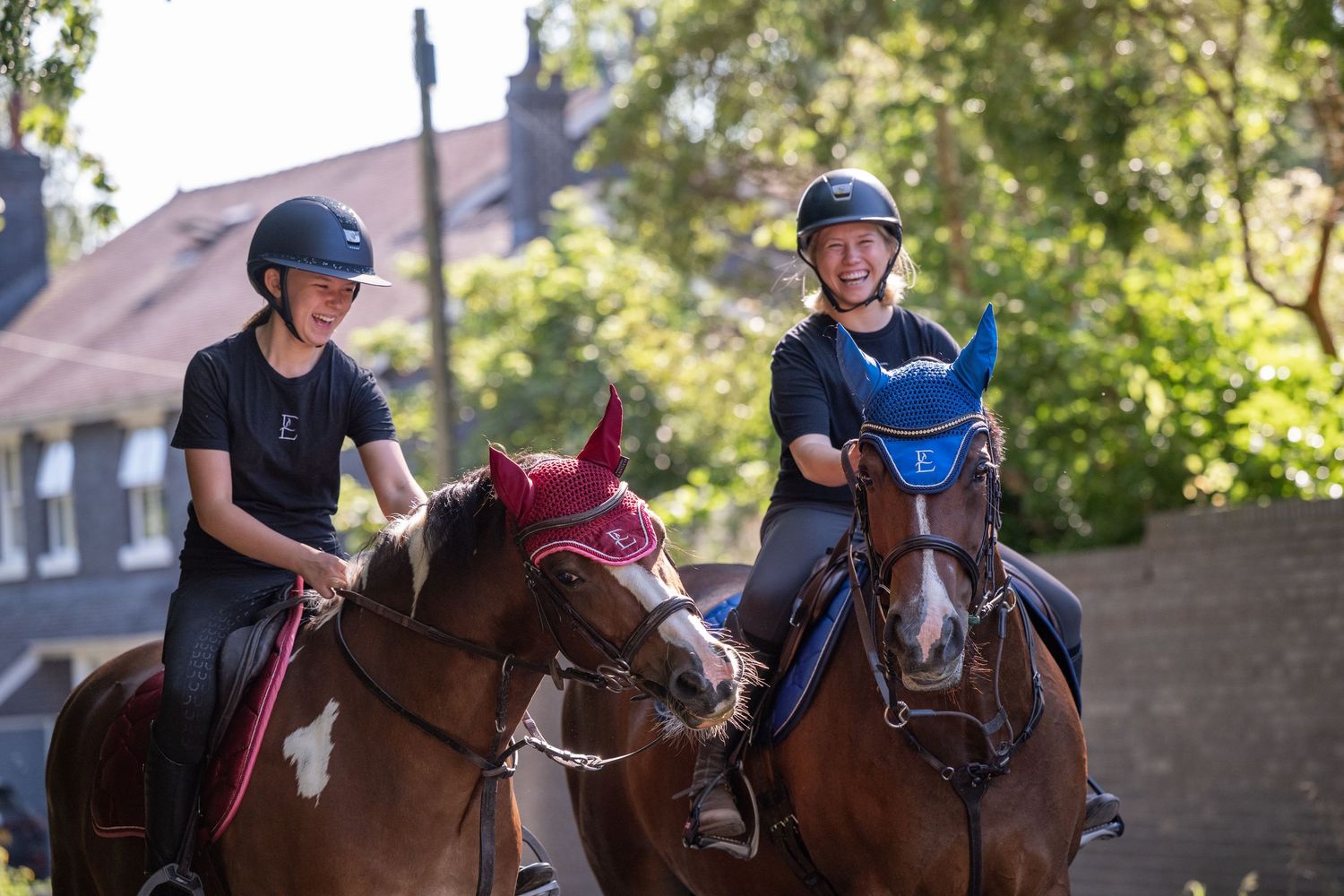Two smiling young riders in helmets on chestnut horses enjoy an outdoor equestrian session on a sunny day.