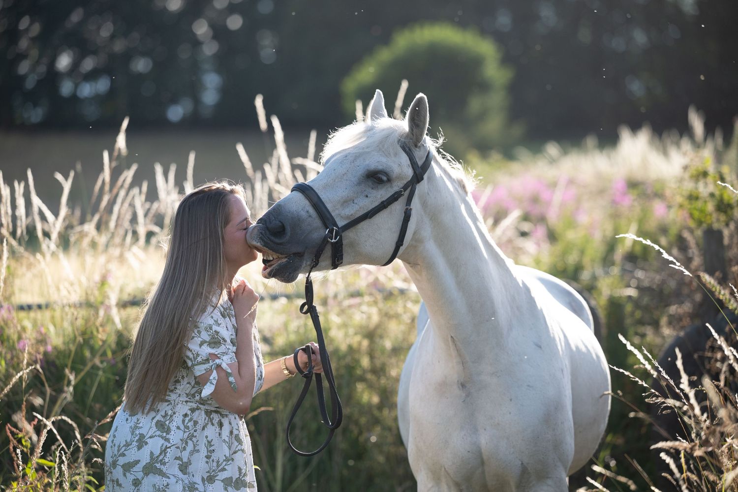 A young woman in a floral dress stands close to a white horse in a sunny meadow.