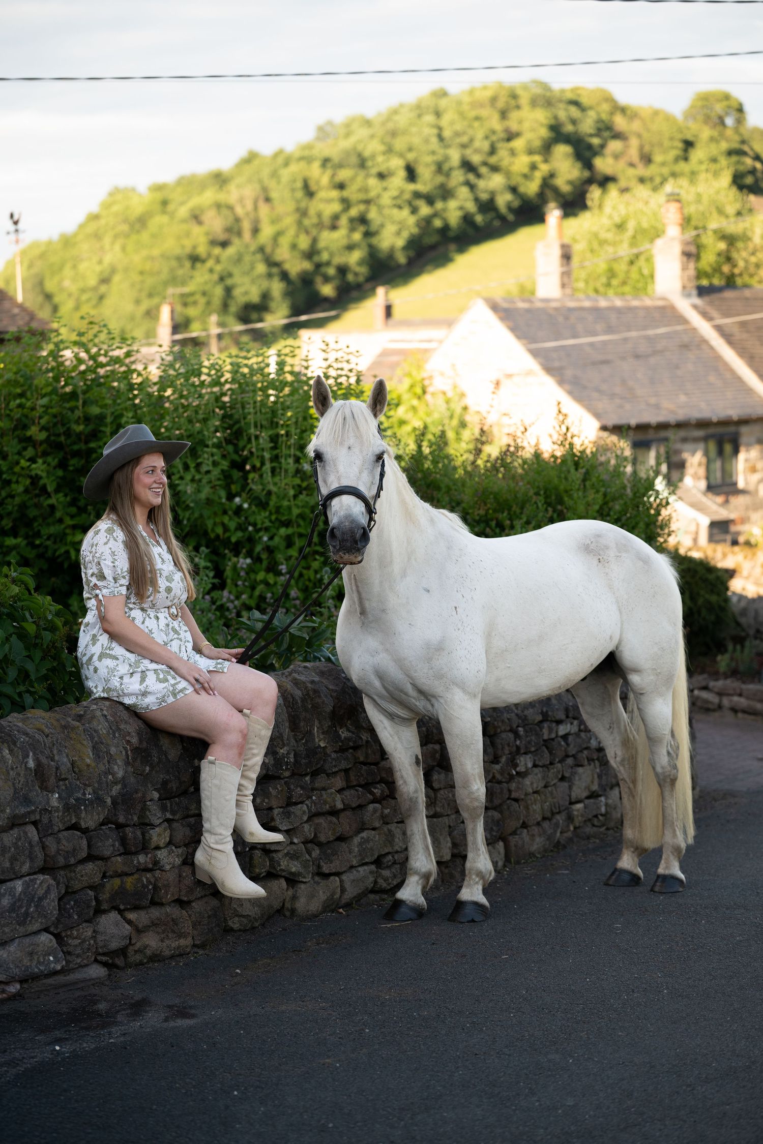 A woman in a floral dress and hat sits on a stone wall beside a white horse in a village.