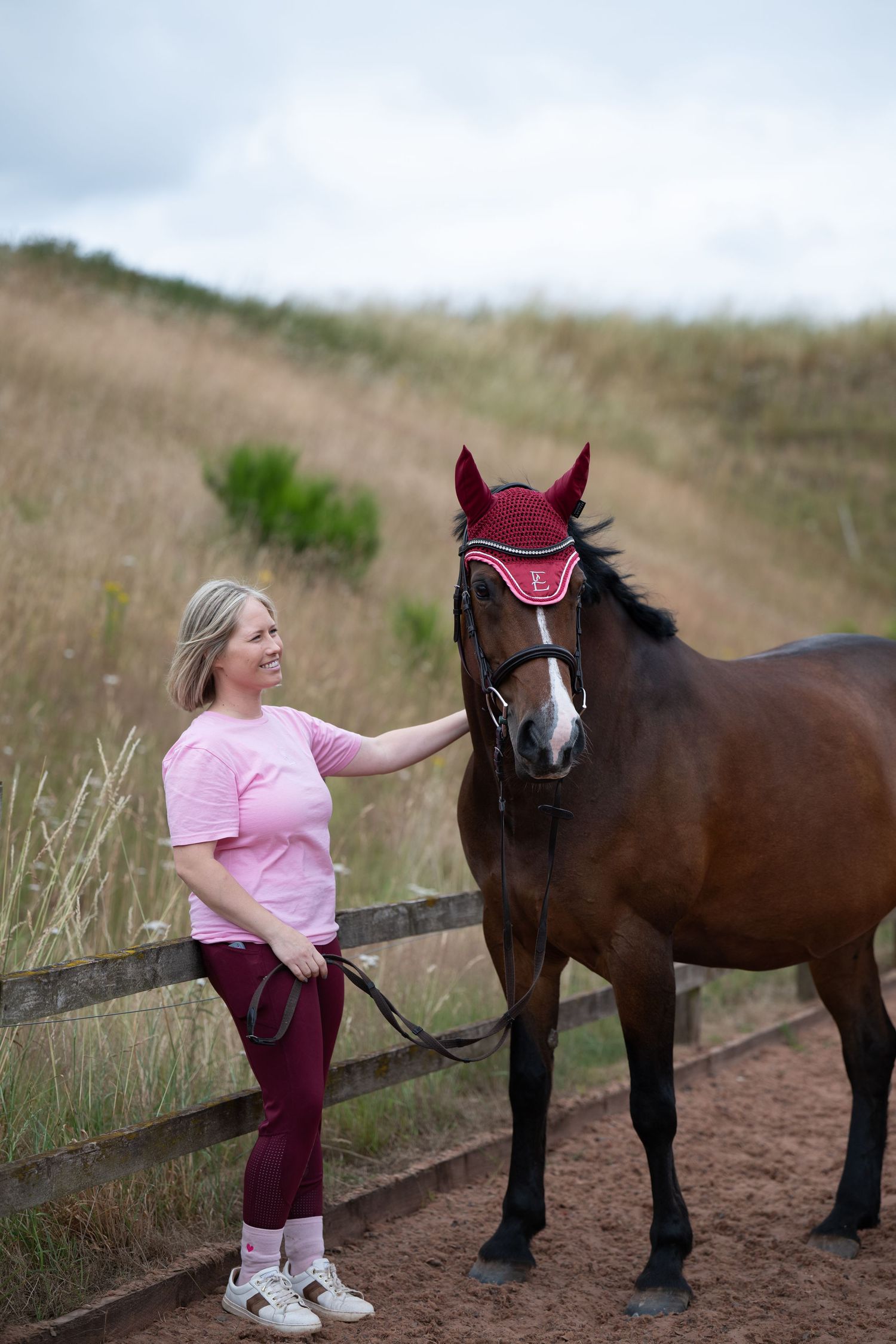 A blonde woman in pink top strokes a bay horse wearing a burgundy fly bonnet outdoors.