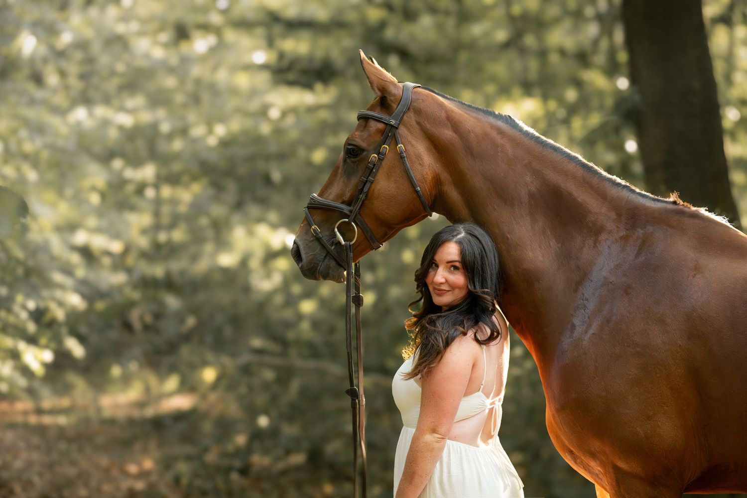 A woman in a white dress stands beside a chestnut horse in a sunlit forest setting.