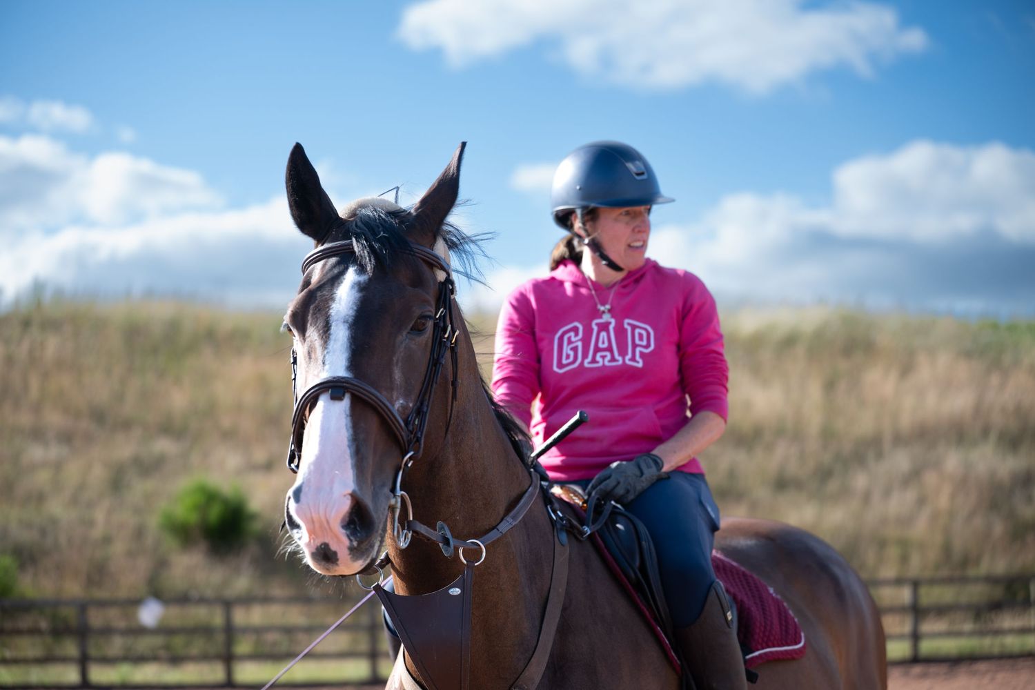 A young girl wearing a GAP hoodie and helmet rides a brown horse in an outdoor equestrian arena.