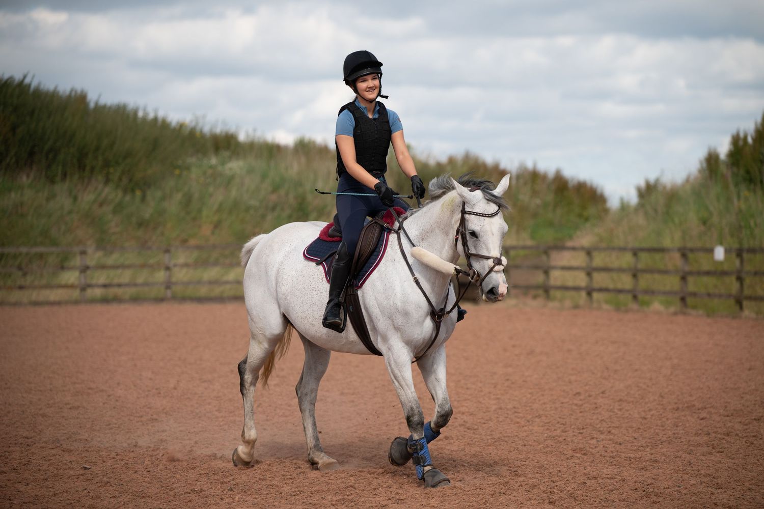 Young rider in helmet on white horse in sandy outdoor arena with wooden fence and grass background.