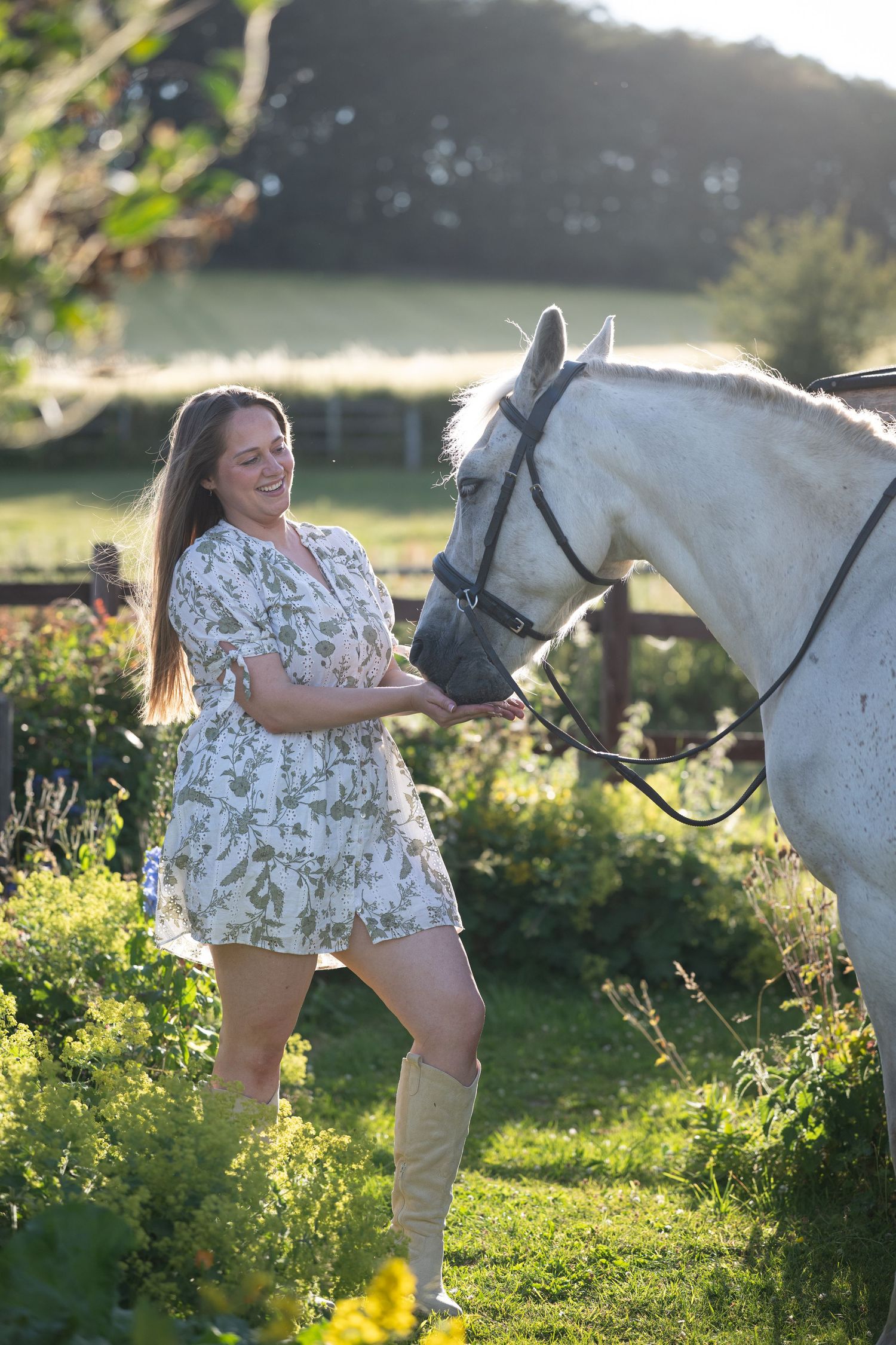 Smiling woman in floral dress and boots feeding a white horse in a sunlit garden.