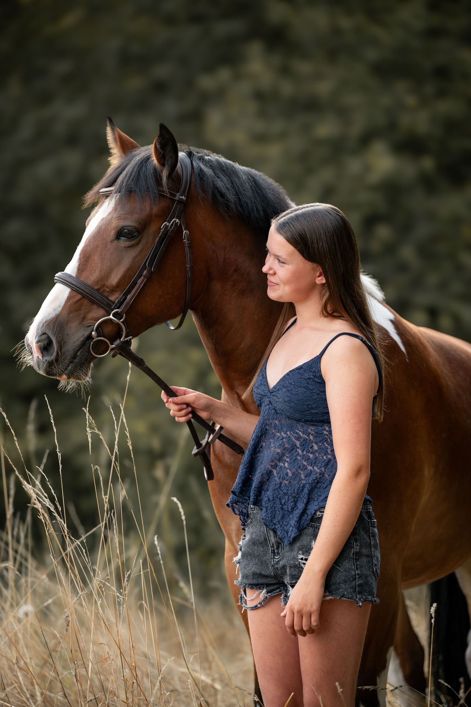 Young girl in denim shorts and blue top stands beside a bay horse in a field of dry grass.
