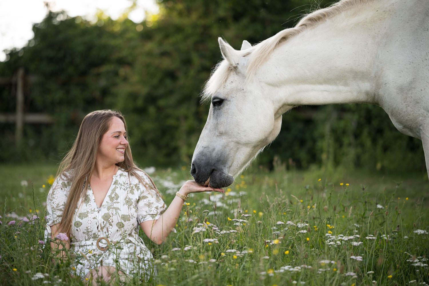 A smiling woman in a floral dress feeds a white horse in a wildflower meadow.