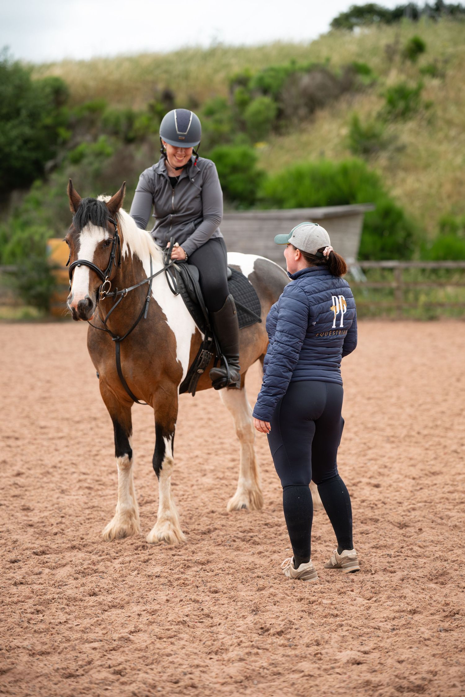 Rider on a brown and white paint horse receiving instruction from a trainer in an outdoor sand arena.