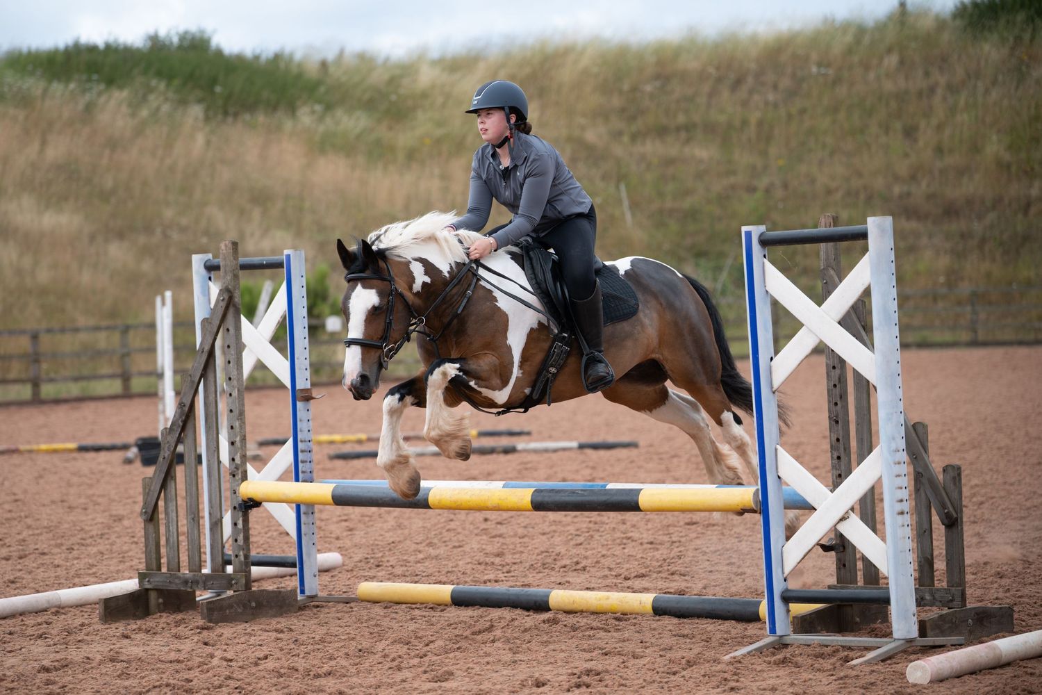 Rider on pinto horse jumping colorful show jump obstacle in sandy equestrian arena during competition.
