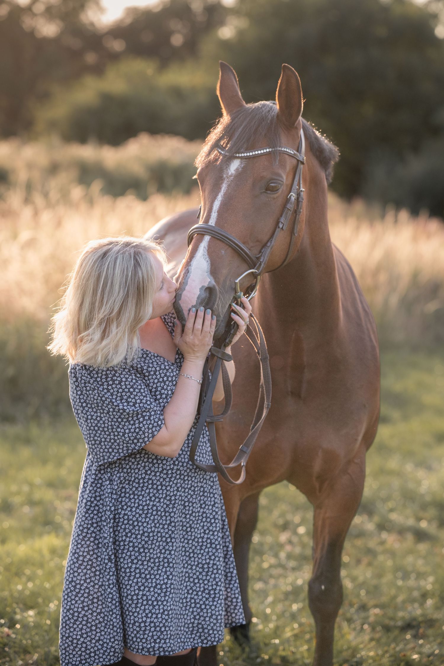 Blonde woman in floral dress affectionately kissing a chestnut horse wearing a bridle in a sunlit field.