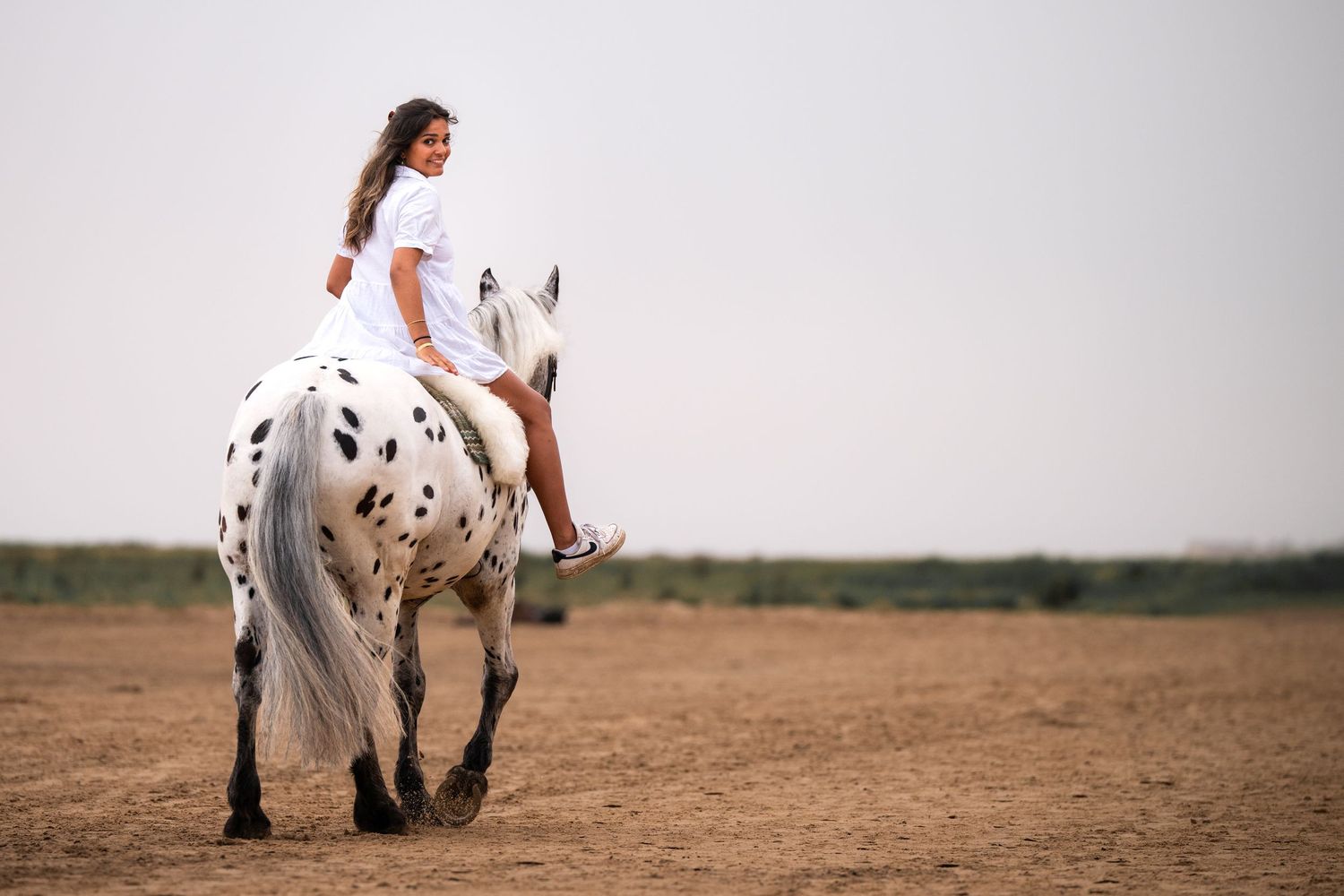 Young woman in white outfit riding a spotted Appaloosa horse on a sandy outdoor arena.