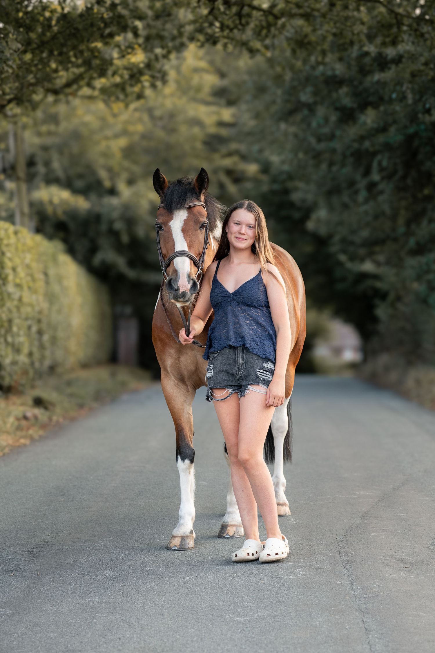Young woman in denim shorts and blue top stands beside a brown and white horse on a tree-lined road.