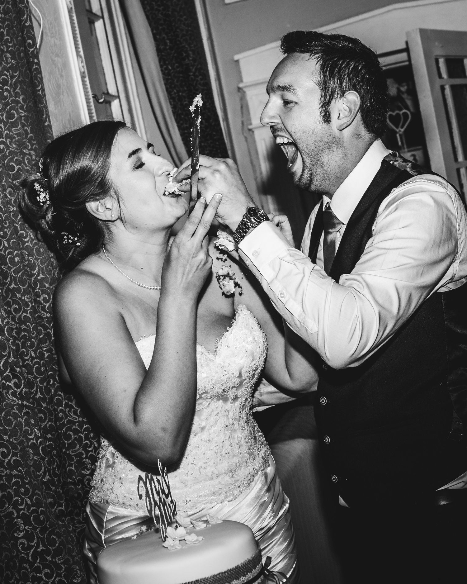 Black and white photo of bride and groom laughing while feeding each other wedding cake at their reception.