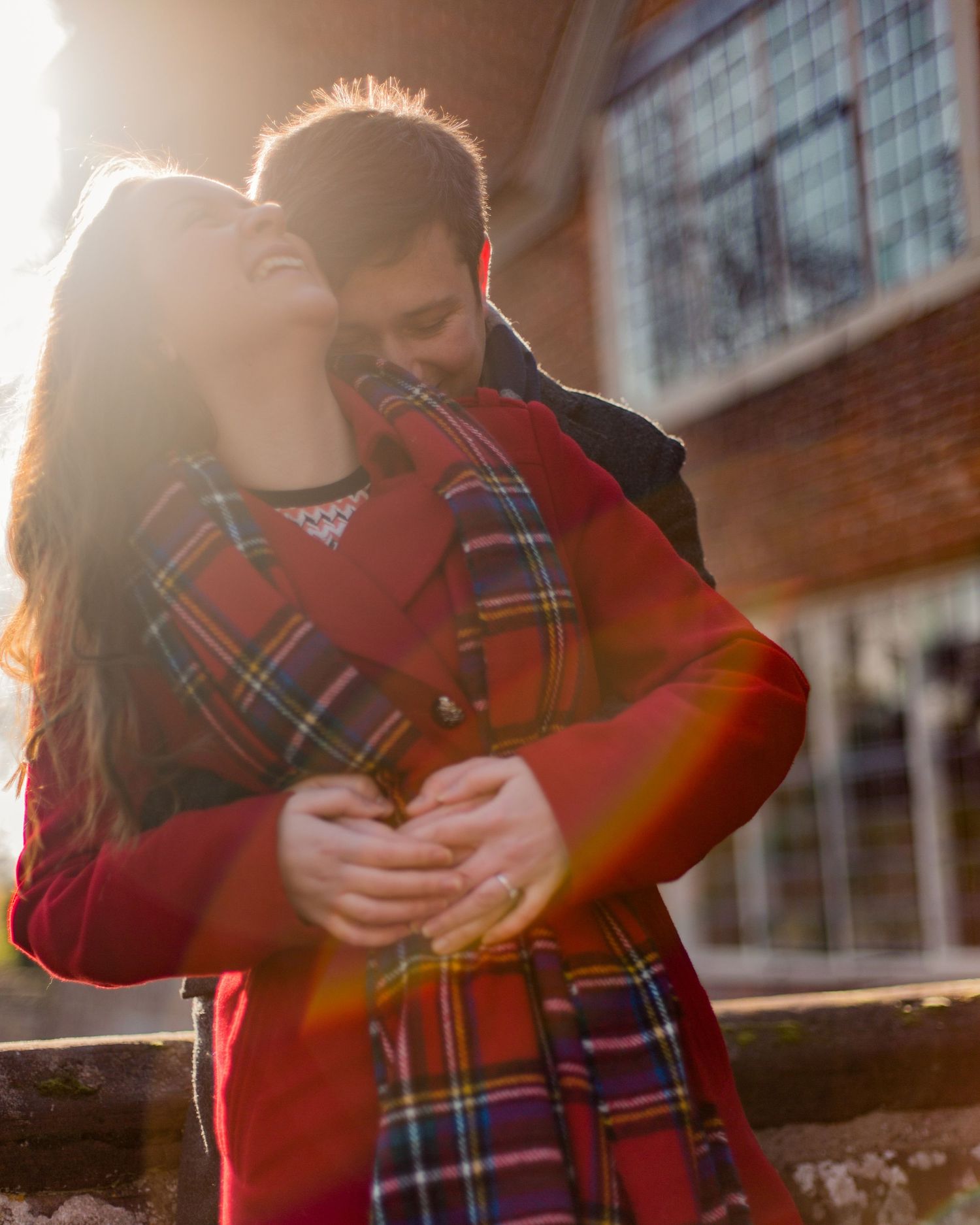 A couple embraces in warm sunset light wearing plaid clothing against an urban backdrop.