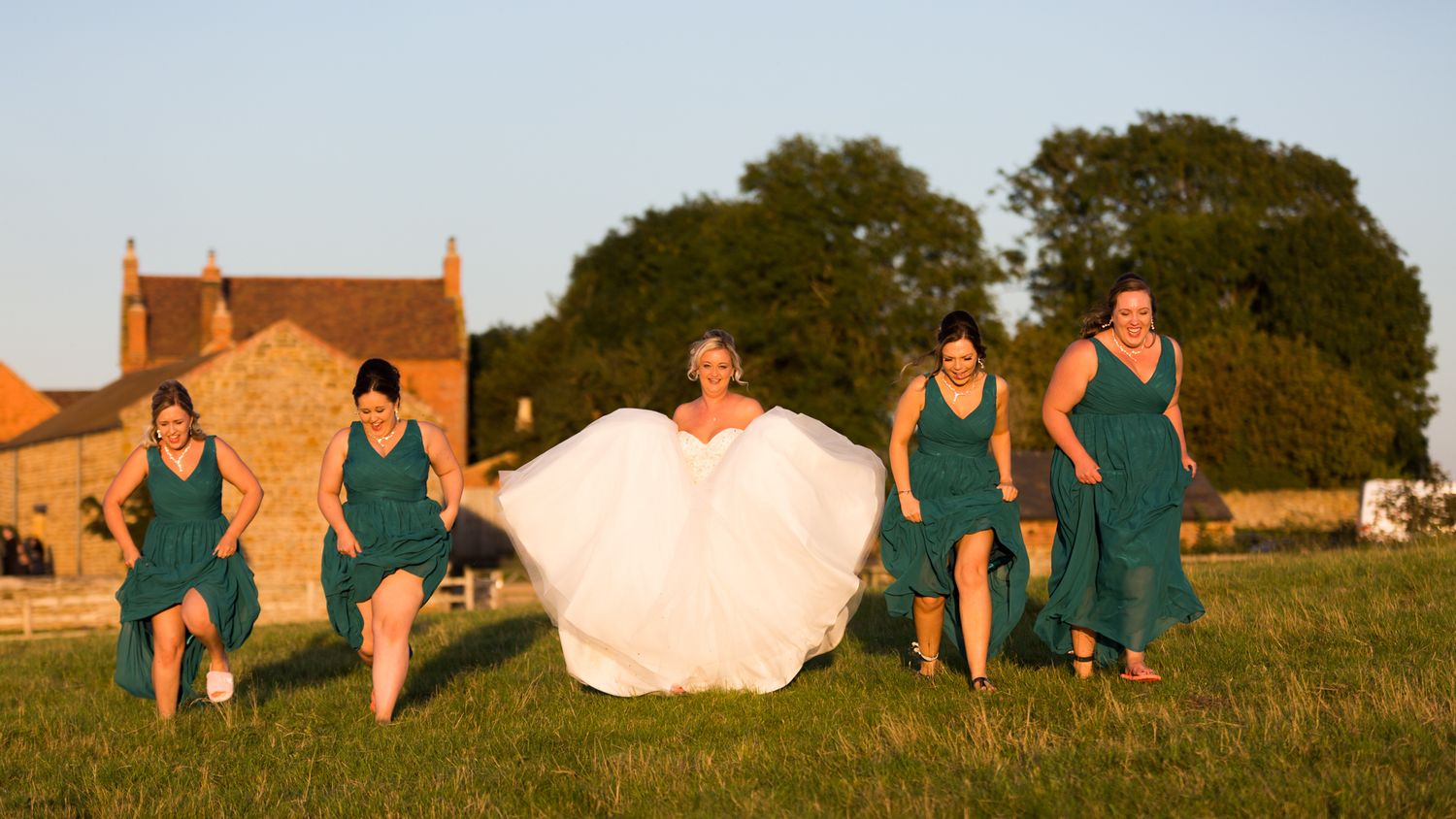 A wedding party in teal dresses runs through a grassy field at sunset with a brick farmhouse in the background.