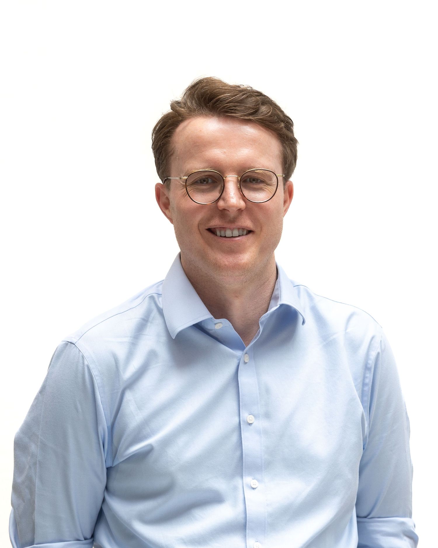 Professional wearing a light blue button-down shirt and round glasses smiles warmly against a white background.