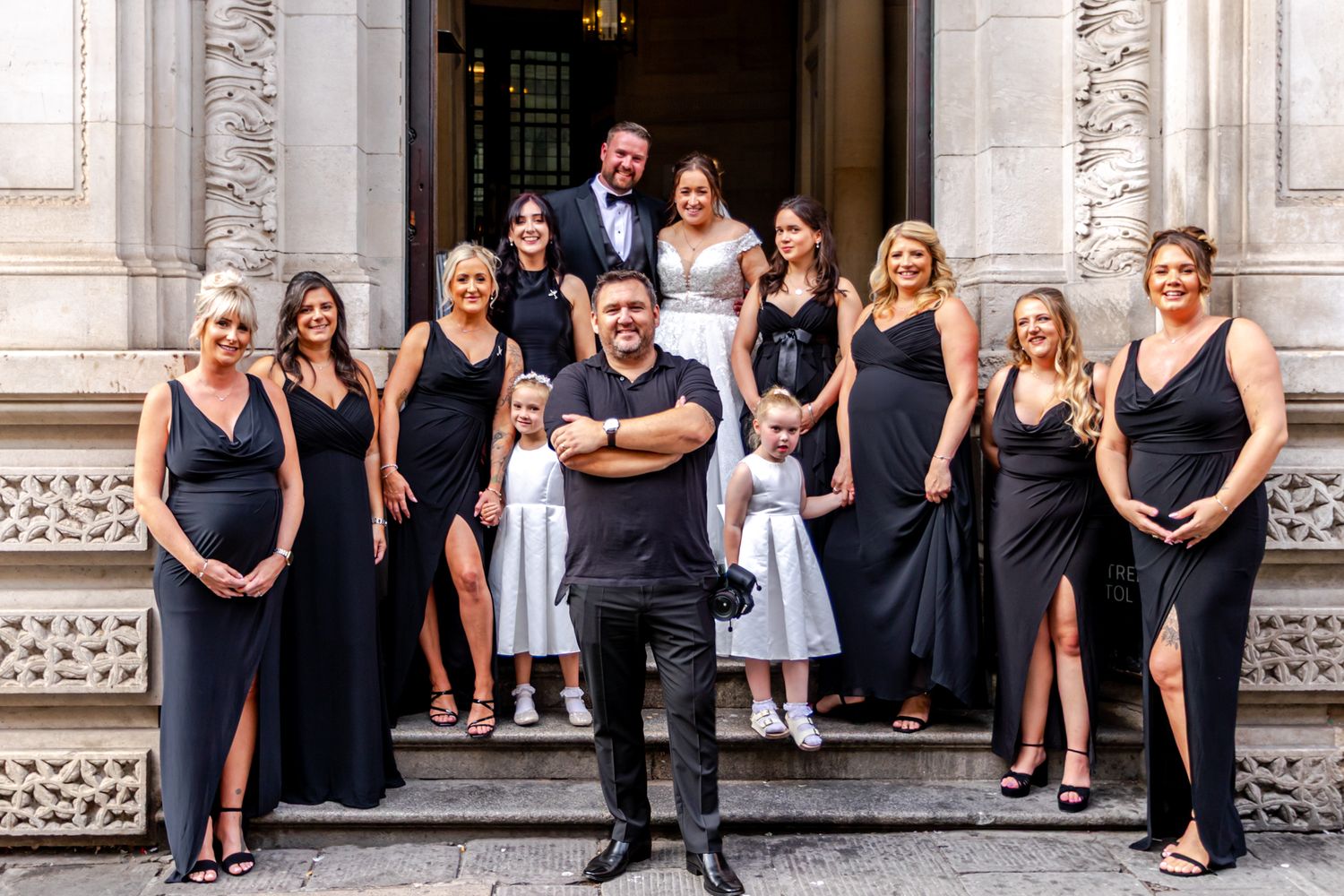 Wedding party in black attire poses on elegant stone steps, bride and groom centered with bridesmaids and flower girls.