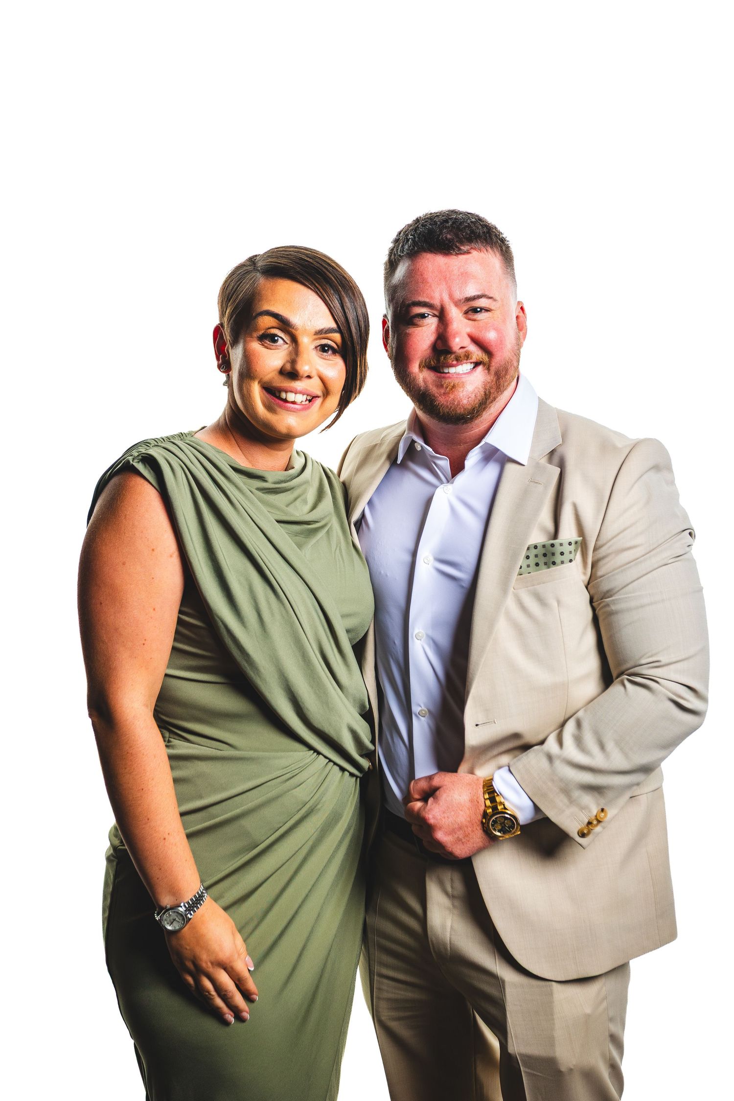 Smiling couple posing together, woman in green dress and man in beige suit with tie, white background.