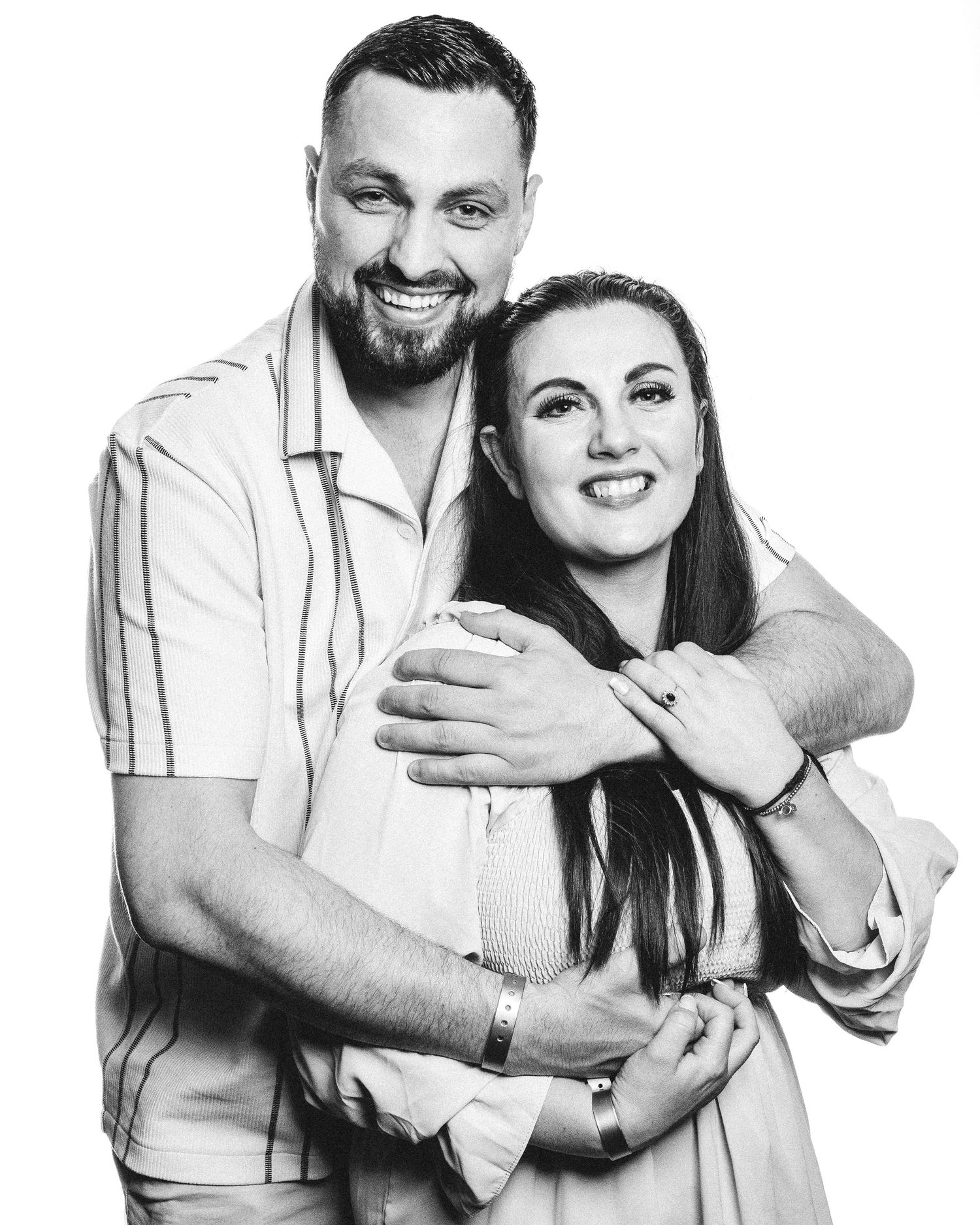 A black and white studio portrait of a smiling couple embracing in light colored outfits against a plain background.