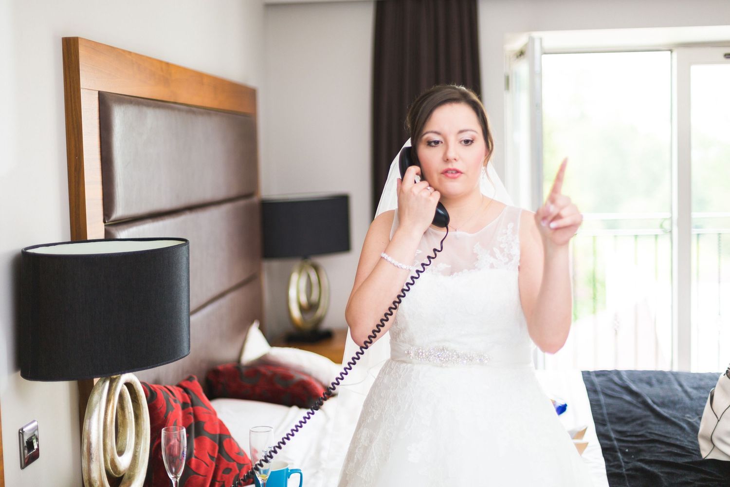 A bride in a white wedding dress holds a landline phone and makes a peace sign while standing in a bedroom.