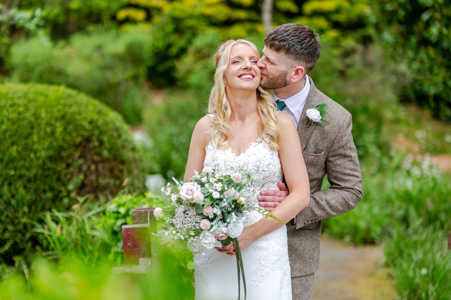 A smiling bride holding a pink bouquet receives a kiss on the cheek from her groom in a lush garden setting.