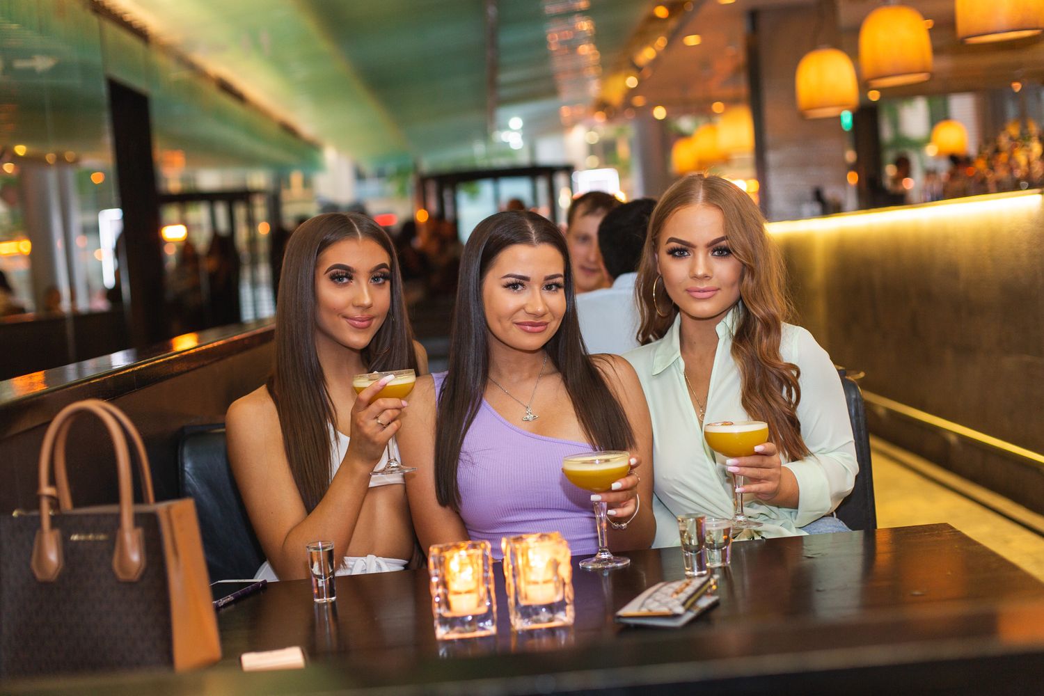 high end wine bar with customers, three young woman dressed in evening wear smiling at the camera