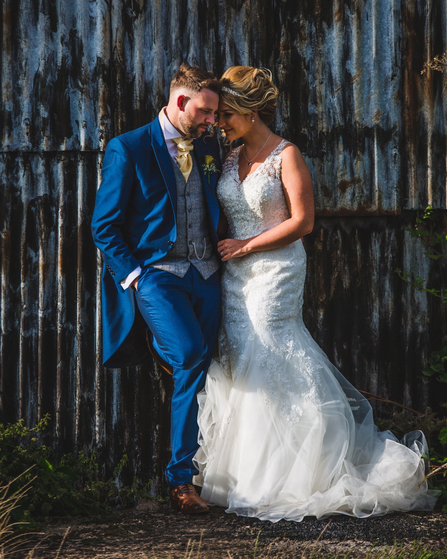 A bride in a white mermaid gown and groom in a blue suit share a romantic moment against a rustic barn wall.