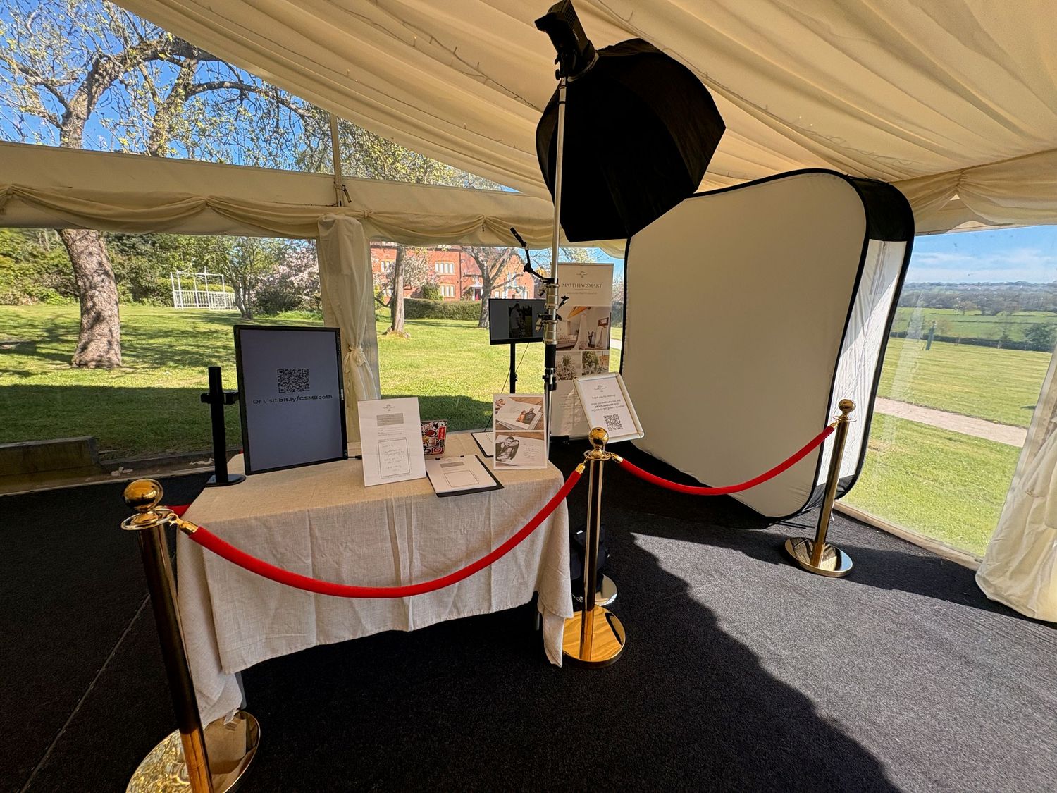 Photo booth setup under a marquee tent with red rope barriers, lighting equipment, and scenic garden views.