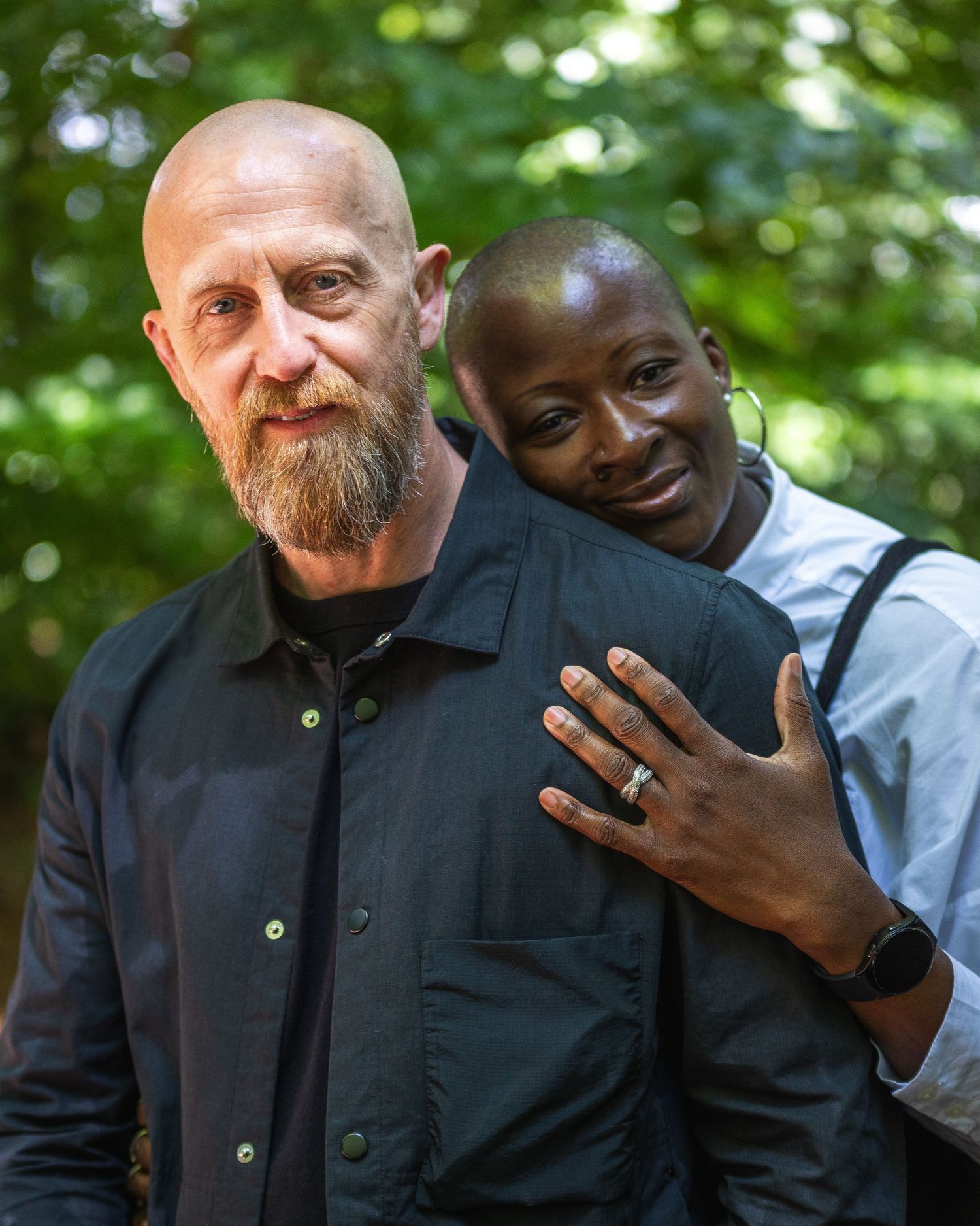 Two people share a tender moment outdoors in front of lush green foliage.