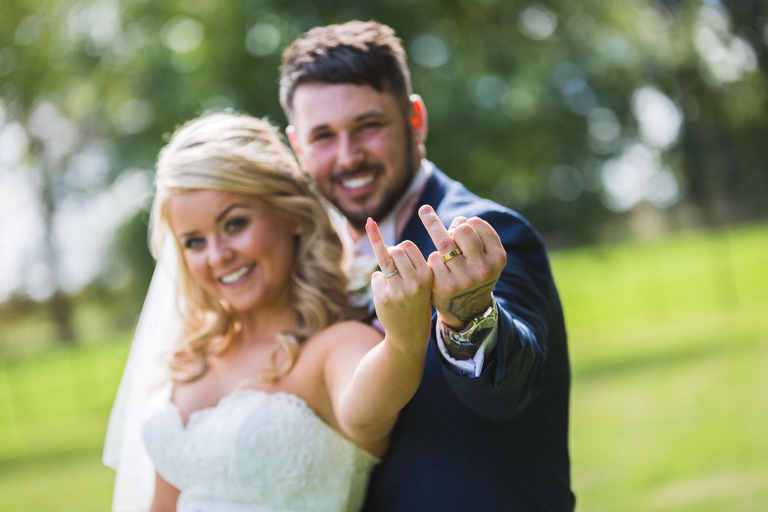 Newlywed couple showing off wedding rings, bride in white dress and groom in suit, smiling outdoors on their wedding day.