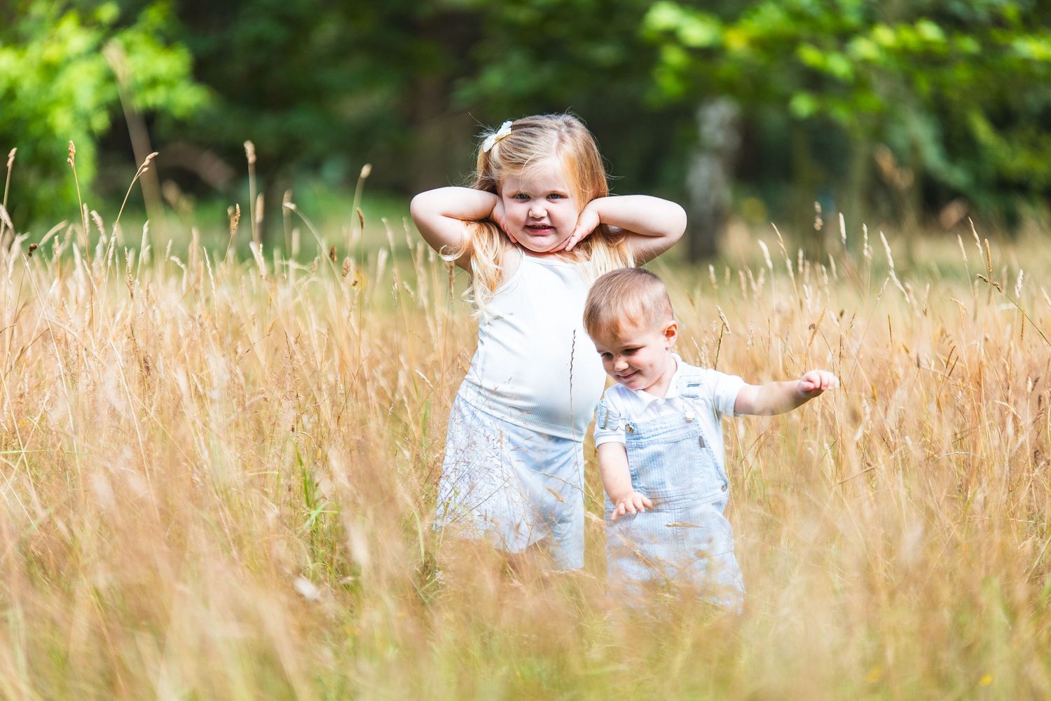Two young children play and pose together while standing in a tall grassy meadow on a sunny summer day.