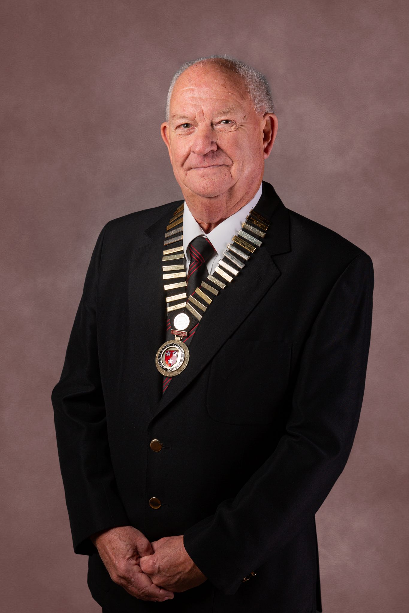 A formal portrait of someone wearing a black suit with a ceremonial chain and medallion against a brown background.