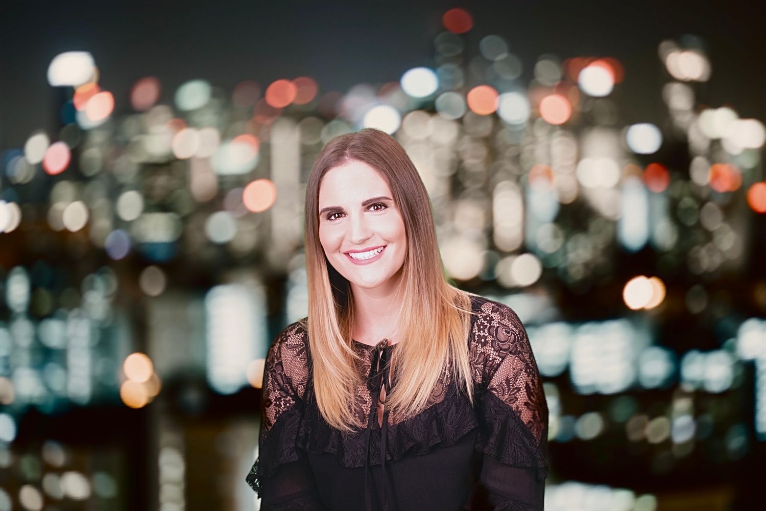 Portrait with blurred city nightscape bokeh lights in background at rooftop location, wearing black lace top.