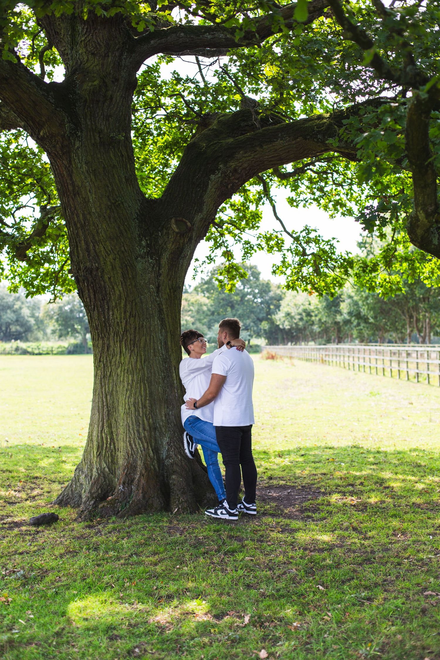 A couple embraces under a large oak tree in a sunny meadow with white fencing in the background.