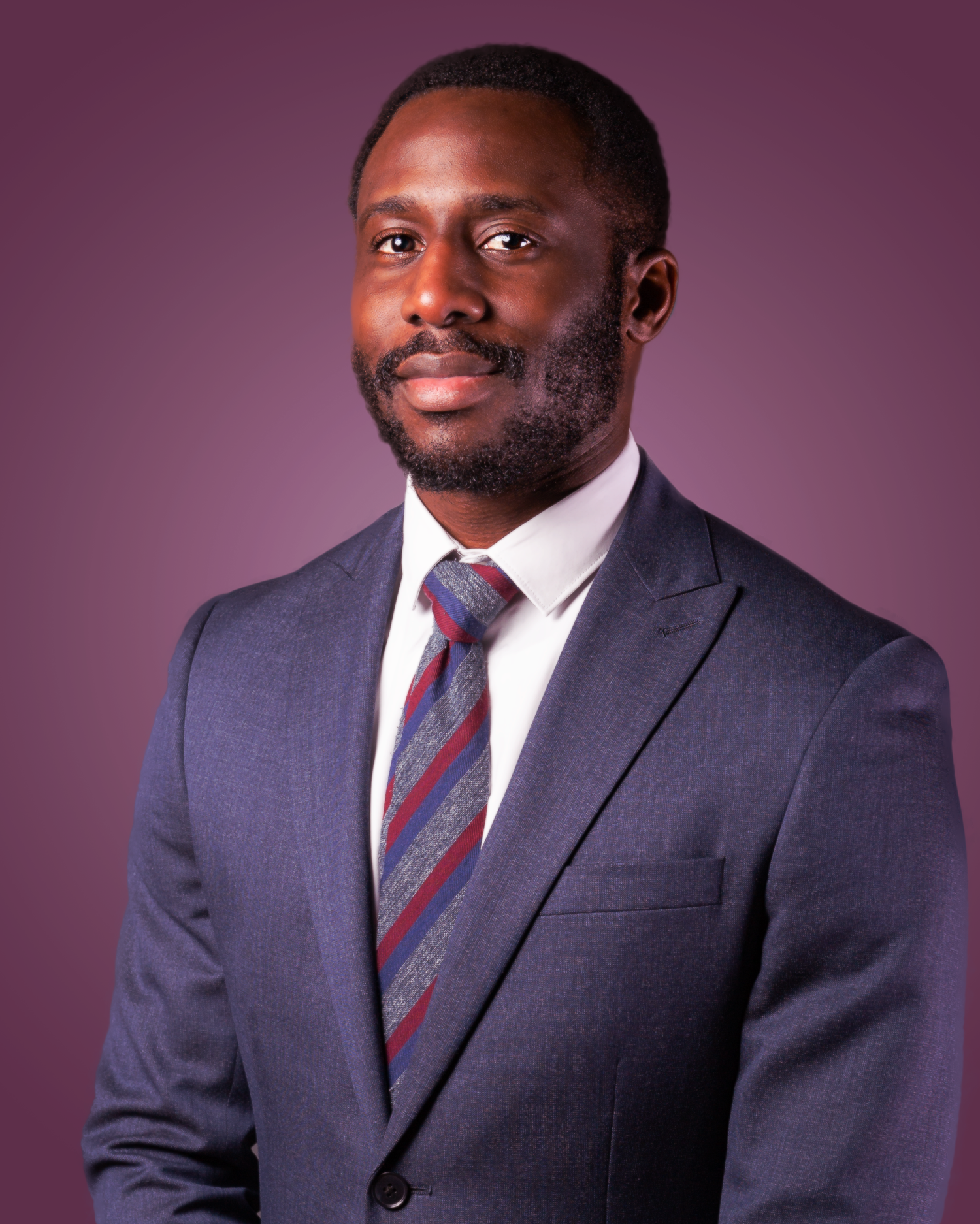 Professional headshot portrait of a person wearing a navy suit with a burgundy and navy striped tie against a purple background.