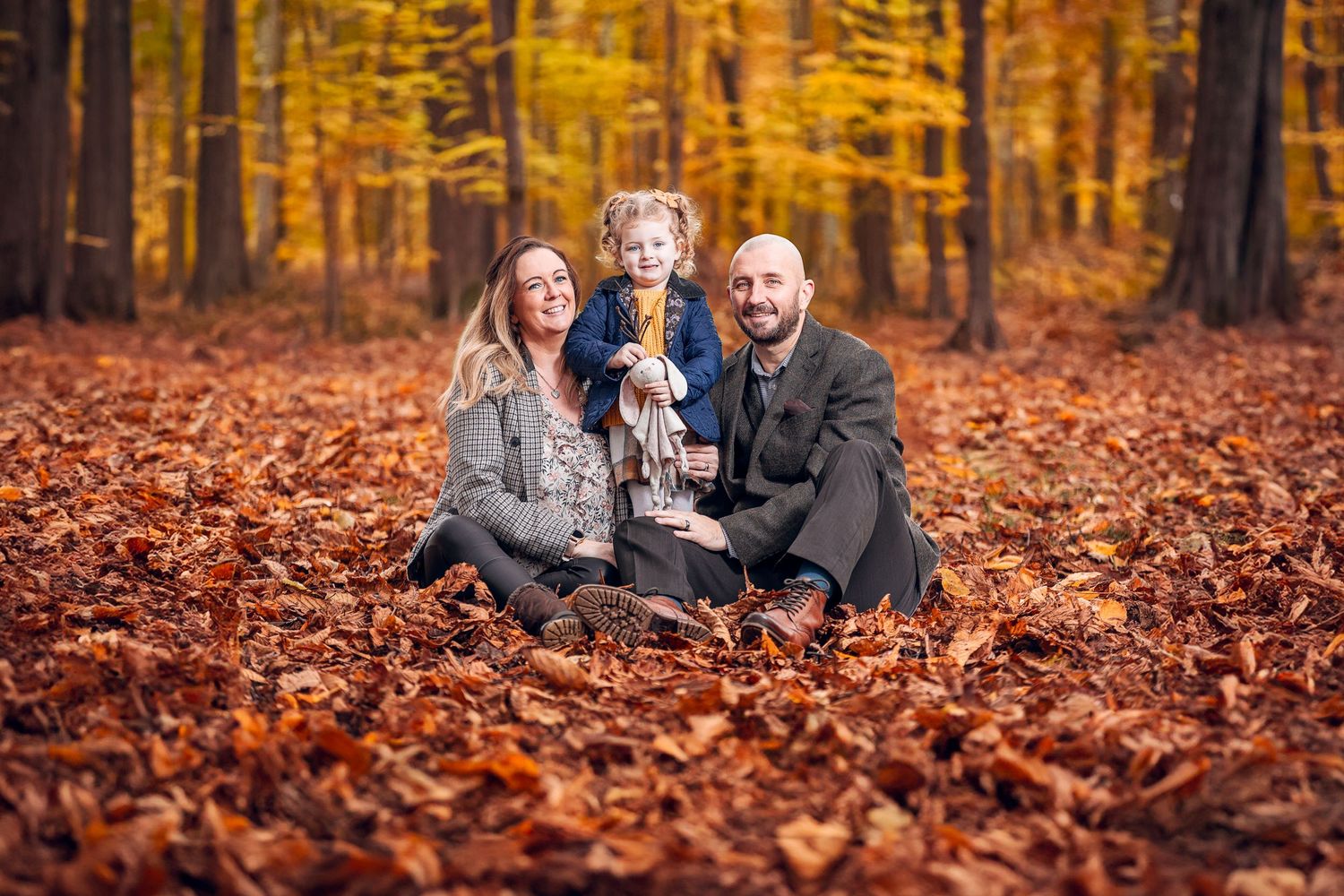 A family sits together on fallen autumn leaves in a forest with vibrant golden foliage in the background.