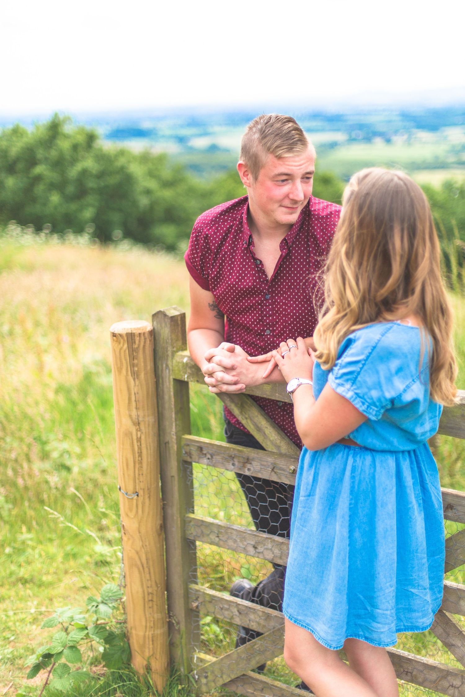 Two people share a sweet moment chatting over a wooden fence in a scenic countryside setting.