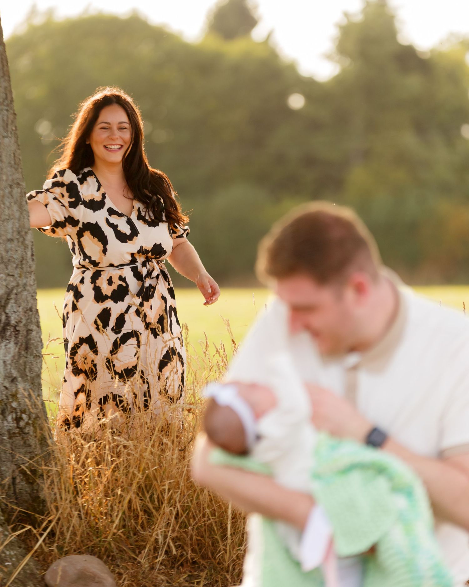 A smiling woman in a floral dress stands by a tree as a man holds a newborn baby in a sunlit field.