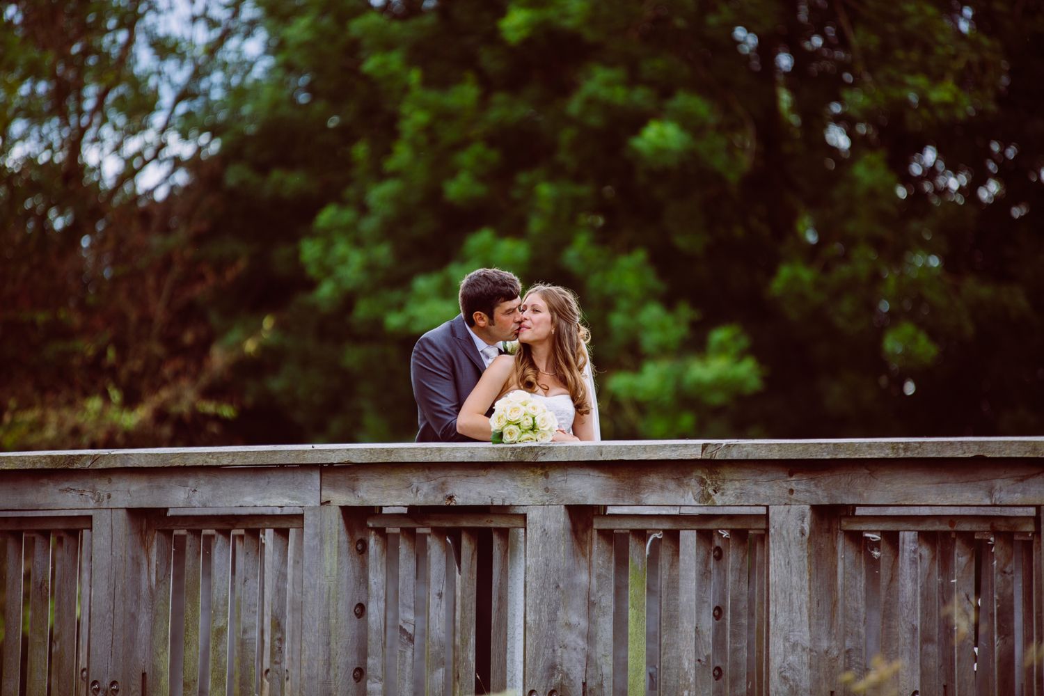 A bride and groom share a romantic kiss on a wooden bridge surrounded by lush green trees on their wedding day.